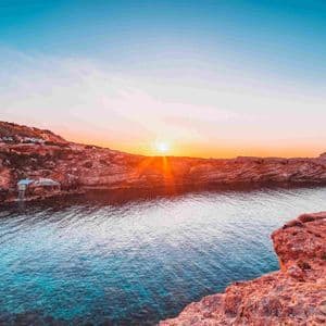 The setting sun casts an orange glow over red rocky cliffs surrounding a calm cove with clear blue water.