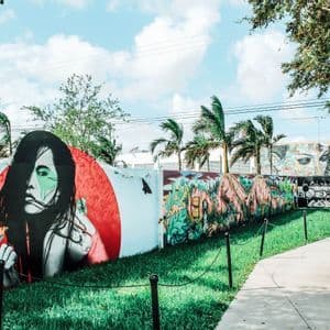 Una parete esterna ricoperta di colorata street art, con un grande murale di una donna, accanto a un prato verde e un sentiero sotto un cielo blu.