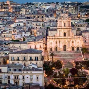 An illuminated baroque church with a grand staircase sits in the middle of a dense hillside city at dusk.