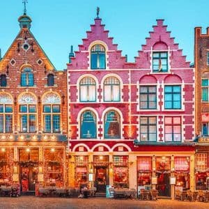 Una hilera de casas de colores, tradicionales y con tejados a dos aguas, decoradas con luces y árboles de Navidad en una plaza adoquinada al anochecer.