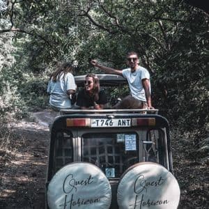 A WeRoad group trip of three people riding in the back of an open-top safari vehicle on a dirt road through a dense forest.