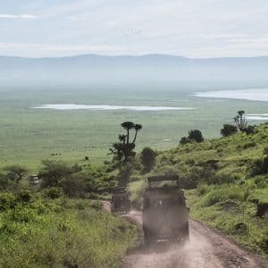 Two safari jeeps drive down a winding dirt road on a grassy hillside, overlooking a vast green valley with a lake and distant mountains.