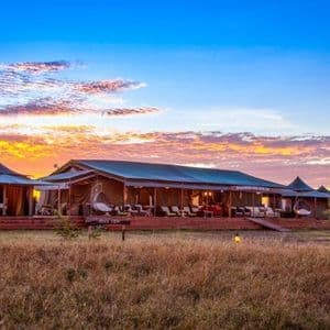 A safari lodge with large tented rooms on a wooden deck, set in a grassy field against a vibrant sunset.