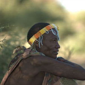 An elderly woman in profile, wearing a traditional beaded headband and cloth, sitting with her arms crossed outdoors.