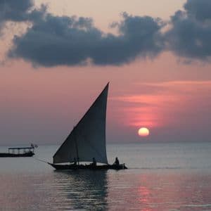 A sailboat with people on board glides across calm water at sunset, with the sun low on the horizon under a pink and cloudy sky.