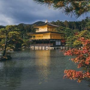 Un tempio giapponese dorato si erge accanto a uno stagno riflettente, circondato da alberi autunnali e colline boscose sotto un cielo nuvoloso.