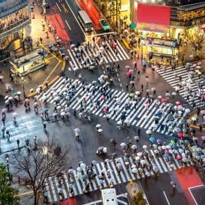Una vista aerea di pedoni con ombrelli che attraversano un incrocio cittadino trafficato in una giornata di pioggia.
