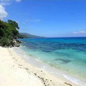 Una spiaggia di sabbia bianca incontaminata si incontra con limpide acque oceaniche turchesi accanto a una costa rocciosa e alberata sotto un cielo azzurro e brillante.