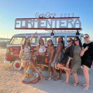 Un viaggio di gruppo WeRoad di donne in posa per una foto davanti a un furgone vintage rosso e bianco su una spiaggia al tramonto.