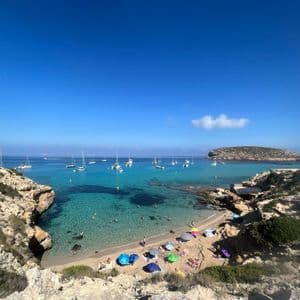 Una vista dall'alto di una caletta sabbiosa con acqua turchese, ombrelloni colorati sulla spiaggia e barche a vela ancorate nella baia.