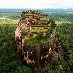 Aerial view of a large rock fortress with ancient ruins on its summit, surrounded by a vast, dense green jungle.