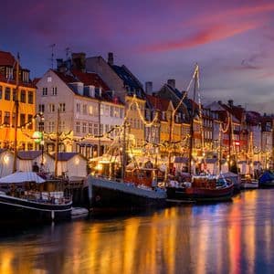 Bunte Ufergebäude und festlich beleuchtete Boote spiegeln sich bei Dämmerung in einem Kanal.