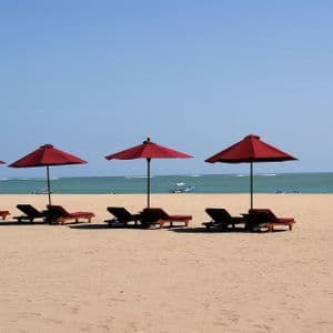 Une rangée de parasols rouges et de transats vides sur une plage de sable fin face à l'océan, sous un ciel dégagé.