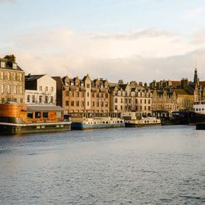 Des bâtiments historiques de la ville et des péniches amarrées longent une rivière paisible sous un ciel nuageux au coucher du soleil.