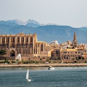 Eine Küstenstadtskyline mit einer großen gotischen Kathedrale, vom Wasser aus gesehen, wo ein Segelboot vorbeigleitet, mit Bergen im Hintergrund.