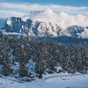 Una fitta foresta di abeti innevati si estende su una collina sotto una grande catena montuosa rocciosa toccata dalle nuvole.