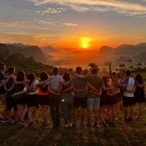 A WeRoad group trip stands with arms around each other, watching the sun set over a misty mountain valley from a grassy viewpoint.