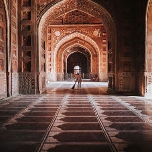 Una persona spazza il pavimento con una scopa in una lunga sala, fiancheggiata da archi intagliati e riccamente decorati, con ombre suggestive.