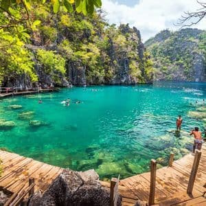 A WeRoad group trip swimming in a clear turquoise lagoon surrounded by rocky cliffs with lush greenery and wooden walkways.
