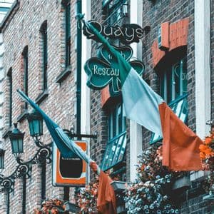 Una bandera irlandesa ondea frente a la fachada de ladrillo de un pub, adornada con coloridas flores y carteles antiguos.
