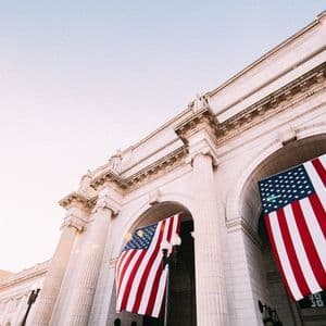 Eine Untersicht auf zwei große amerikanische Flaggen, die von den Bögen eines klassischen Steingebäudes herabhängen.
