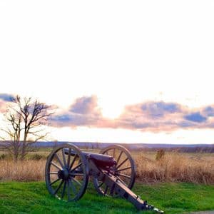 Eine historische Kanone steht auf einem grasbewachsenen Hügel und überblickt ein Feld mit einem kahlen Baum vor einem bewölkten Sonnenuntergangshimmel.