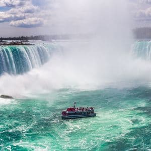 Ein Ausflugsboot mit einer WeRoad-Gruppe navigiert durch die nebligen, türkisfarbenen Gewässer am Fuße eines riesigen, gekrümmten Wasserfalls.