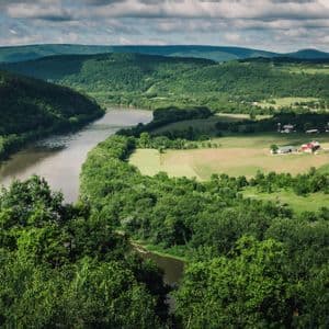 An elevated view of a river winding through a valley of green forests and fields, with rolling hills in the distance under a cloudy sky.