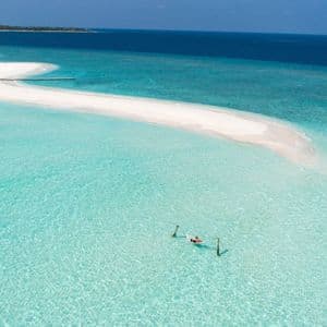 Vista aerea di una persona che galleggia su una zattera in acque cristalline turchesi, accanto a un sinuoso banco di sabbia bianco e all'oceano blu profondo.