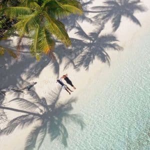 An aerial view of two people relaxing on a white sand beach next to clear water, with shadows from palm trees.