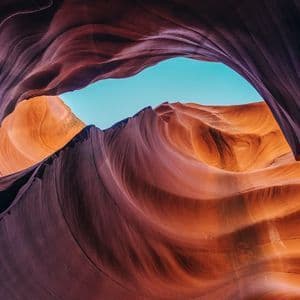 Wavy, layered sandstone formations of a slot canyon in orange and purple tones, framing a patch of clear blue sky.