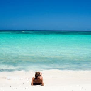Una donna con i capelli raccolti a chignon giace su una spiaggia di sabbia bianca, guardando il mare calmo e turchese.