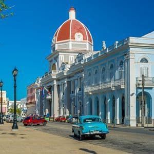 Un coche azul clásico circula por una calle junto a un gran edificio con una cúpula roja bajo un cielo azul claro.