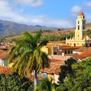 Un campanile giallo si erge su una città con tetti in terracotta, con palme e montagne verdi sullo sfondo.