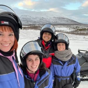 Vier Frauen einer WeRoad-Gruppenreise mit Helmen lächeln für ein Selfie neben einem Schneemobil in verschneiter Berglandschaft.