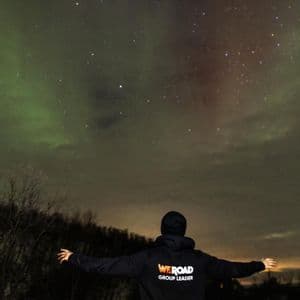 Ein WeRoad-Reiseleiter steht mit ausgestreckten Armen da und blickt zum Polarlicht und zum sternenklaren Nachthimmel auf.