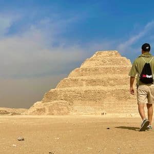 Un viaggiatore con uno zaino WeRoad attraversa un paesaggio desertico sabbioso verso una grande piramide a gradoni.
