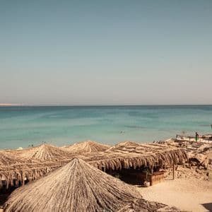 Strohschirme an einem Sandstrand mit zwei Booten, die in ruhigem, türkisfarbenem Wasser unter klarem Himmel ankern.
