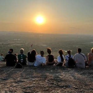 A WeRoad group trip sits on a rocky outcrop, watching the sun set over a sprawling green valley and distant hills.
