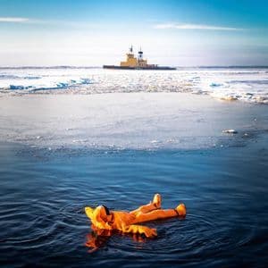 Une personne en combinaison de survie orange flotte sur le dos dans l'eau glacée, avec un brise-glace au loin.