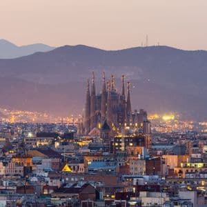 Un paysage urbain illuminé avec une grande cathédrale ornée et une chaîne de montagnes en arrière-plan, au crépuscule.