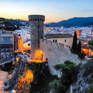 Eine Steinburg mit einem Turm liegt beleuchtet auf einer Klippe mit Blick auf eine Stadt und Berge bei Sonnenuntergang.