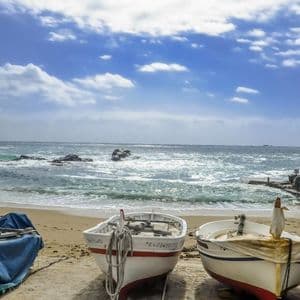 De petits bateaux de pêche reposent sur une plage de sable avec une mer agitée et un ciel nuageux en arrière-plan.