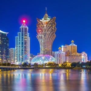 Grattacieli illuminati costeggiano un lungomare di notte, con un ponte sulla sinistra e luci colorate che si riflettono sull'acqua.