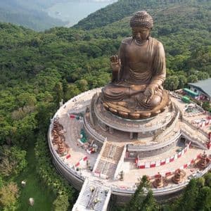 Veduta aerea di una grande statua di Buddha in bronzo, situata in cima a una piattaforma a più livelli su una montagna coperta da una fitta foresta verde.