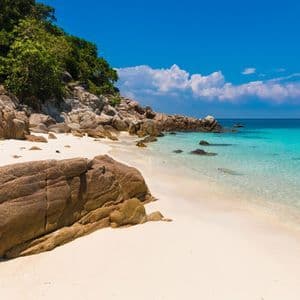 Une plage de sable blanc isolée, parsemée de grands rochers, bordée d'une végétation luxuriante et baignée par des eaux calmes et turquoise sous un ciel bleu.