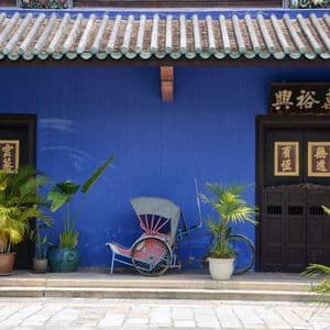 Un rickshaw tradicional y plantas en maceta se encuentran frente a un edificio azul vibrante con puertas oscuras y un tejado de tejas.