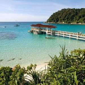 Un pontile di legno si estende attraverso una baia tranquilla, chiara e turchese verso una lussureggiante collina verde in una giornata di sole.