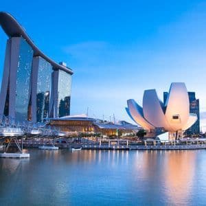 Uno skyline moderno di città al crepuscolo, con monumenti architettonici illuminati come un edificio a forma di loto e un grattacielo con una nave in cima, riflessi nell'acqua.