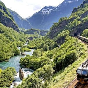 Un treno viaggia su un binario attraverso una lussureggiante e verde valle di montagna accanto a un fiume, con cime innevate in lontananza.